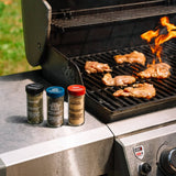 Three spice seasoning bottles labeled Pepper Jack, Garlic Pepper, and Garlic Powder sit on the side of a stainless steel grill with chicken cooking on the grate and flames visible.