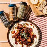 Three spice jars labeled Whole Black Pepper, Ground Cinnamon, and Rosemary are arranged around a wooden board holding a spread of white dip with glazed nuts, pecans, and a rosemary sprig, with sliced bread on a striped cloth in a warm styled setting.