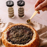 A round bread bowl filled with dark jam and topped with rosemary, set on a beige cloth. Two spice jars labeled Rosemary and Thyme stand behind it, and a hand holds a small gold spoon sprinkling seasoning.