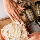Hands spreading a thick herb butter mixture onto a raw ground meat loaf, with several bottles of Marie Rosemary seasoning visible on a striped cloth and baking sheet.