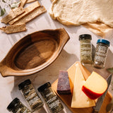 An overhead styled food scene on a light stone surface featuring a rustic brown ceramic bowl, sliced cheeses, crackers, herbs, and several spice jars. No person or animal is present, but this is not a clean isolated product shot of a single sellable item; it reads as a lifestyle/food-styling composition, so subject_type is Lifestyle.