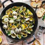 Skillet of clams with lemon wedges and herbs on a wooden table with bread slices.