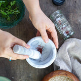 A top-down view of hands using a white marble mortar and pestle to grind red saffron threads in a white marble bowl, with a jar of saffron threads, a green bowl of cilantro, and a beige cloth on a dark wooden table.