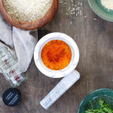 A white marble mortar and pestle filled with orange-red ground spice, arranged on a rustic wooden table with bowls of rice, herbs, and a bottle of saffron threads nearby.