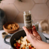 A hand holds a clear glass spice jar labeled 'SAGE' and 'Cayenne & Bassette' in front of a kitchen scene with a pot of chopped vegetables.