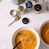 Two bowls of orange-colored soup topped with herbs and nuts, arranged on a light stone surface with several spice jars and a small spoon of seasoning nearby.