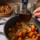 A close-up of a hand pouring amber liquid seasoning from a metal measuring cup into a dark gray pot containing sautéed onions, carrots, and potatoes. Several spice jars and a wooden bowl of nuts are blurred in the background on a light countertop.