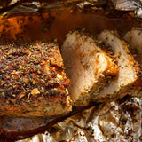 Close-up of a roasted meat dish, showing a sliced piece with a heavily seasoned browned crust and pale pink interior on crinkled foil.