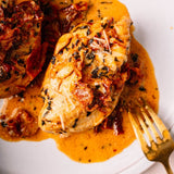 Close-up of a plated cooked chicken breast in a rich orange sauce with sunflower seeds and herbs, with a gold fork on a white plate.