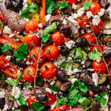 A close-up overhead shot of a colorful Mediterranean salad filled with cherry tomatoes on the vine, crumbled feta cheese, fresh parsley, black olives, zucchini, red onion, and roasted peppers.