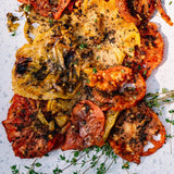 A top-down close-up of a mixed vegetable salad on a speckled white plate, featuring roasted red and yellow tomatoes, caramelized onions, and fresh thyme sprigs sprinkled with black pepper.