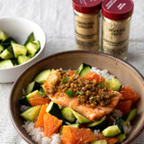A prepared salmon bowl over white rice with cucumber, orange segments, and a brown sauce, with two spice jars labeled Ground Ginger and Sesame Seed standing behind it on a light textured surface.