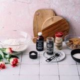 A styled flat-lay of baking ingredients and wooden cutting boards on a white tiled surface against a pink wall. Visible items include a glass bowl of flour, red tulips, a bottle of vanilla extract, a jar of black seed, a jar of sesame seed, a bowl of brown sugar, and a bowl of white sugar.