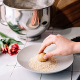 A hand is dipping a small round cookie into sesame seeds on a light gray plate, with a stainless steel mixing bowl and tulips in the softly blurred background.