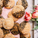 A top-down view of several round sesame-topped pastries arranged on a light pink cloth over a white wooden board, with pink tulips on the right side.