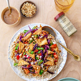A styled overhead shot of a prepared noodle dish on a speckled white plate, with tofu, purple cabbage, red bell peppers, green beans, and sesame seeds. Surrounding the plate are a small bowl of peanut sauce, a bowl of crushed peanuts, a glass of amber liquid, and a jar labeled Sesame Seed.