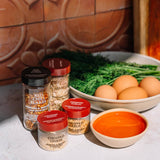 Four jars of Morton's seasoned rice flakes arranged on a white marble countertop with cooking ingredients, including a bowl of eggs, fresh dill, and a bowl of orange seasoning mix, against a rustic terracotta tiled background.