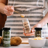 A person in a gray and white striped shirt holds a clear glass jar of Shalott Morton & Co Asparagus & Garlic Seasoning, lifting a spoonful with a gold spoon. Other seasoning jars labeled Chive, Parsley, and Basil are arranged on a table with a potato and a bowl of butter in the foreground.