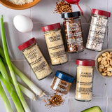 Five spice jars arranged on a light tiled surface with food props including green onions, peanuts, rice, garlic, and a red radish. The jars are labeled Morton & Bassett spices: toasted sesame seed, ground ginger, red chili flakes, shalots, and garlic powder.