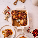 Overhead food styling shot of baked chicken in a white rectangular baking dish with a fork, alongside two spice jars, plates with portions, cranberries, and a cutting board on a light stone surface.
