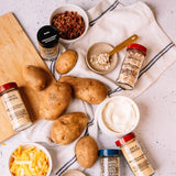 An overhead flat-lay of various gourmet seasoning bottles, bowls of chopped ingredients including shredded cheese and cooked onions, and several whole russet potatoes on a white cloth over a light stone surface.