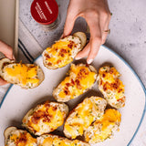 Overhead view of baked potato halves topped with melted cheese and crispy bacon bits, arranged on a speckled white plate. Two hands are visible holding portions of the food, with a bottle of seasoning in the background on a light stone surface.