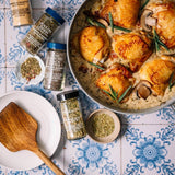 A styled overhead flat-lay of a cooked chicken and mushroom dish in a pan, surrounded by several spice jars and small bowls of seasoning on a blue-and-white patterned tile surface. Visible spices include smoked paprika, garlic powder, ground black pepper, and herb blends.