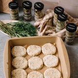 Nine raw round fishcakes or patties sit in a beige baking pan lined with parchment paper, with several labeled spice jars and fresh rosemary sprigs arranged around it on a countertop.