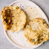 Two halves of a baked bread roll or biscuit with a golden-brown, textured crumb, served on a speckled off-white ceramic plate against a light gray background.