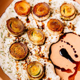 A close-up overhead shot of a flatbread pizza topped with sliced caramelized onions, herbs, and a peach-colored sauce drizzled with dark balsamic glaze.