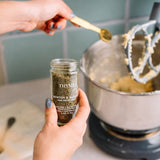 A person holds a small glass jar of Morton & Bradley San Francisco Thyme Seasoning in a kitchen setting, with a measuring spoon adding seasoning to a stand mixer bowl in the background.