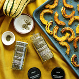 Two glass spice jars labeled Thyme and Sage with black lids, arranged on a yellow fabric next to roasted squash on a baking tray, with small bowls of herb seasoning.