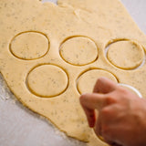 A hand using a round cutter to cut circles from a rolled sheet of speckled dough on a light surface.
