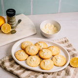A white oval platter holding several round golden cookies topped with shredded cheese, set on a beige checkered cloth. A small bowl of cheese, a jar of dried thyme, and lemon slices are arranged on a white cutting board against a pale blue tiled background.