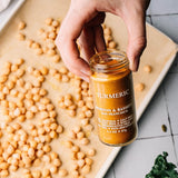 A hand holds a glass jar of Morton & Bassetty Turmeric San Francisco seasoning over a baking sheet filled with chickpeas.
