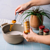 A person's hands are shown holding a glass jar of Turmeric & Hopsi Spice Blend and sprinkling orange-yellow powder into a beige ceramic bowl. The background features a potted plant and other spice jars on a light blue tiled surface.