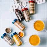 An overhead flat-lay of several spice jars and small bowls of ground spices arranged on a light blue tiled surface, with two white mugs of orange-yellow curry. A striped cloth is visible at the top.