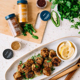 A styled overhead food scene showing prepared meatballs on a long white platter with lemon slices in a small bowl, alongside spice jars and herbs on a cutting board and countertop.