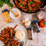 A styled flat-lay of two spice blends, Turmeric and Ground Chipotle Pepper, arranged on a light stone tile surface with cooked sweet potato fries, a plate of rice, garlic, onion, and a tomato.