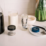 Two small round cosmetic jars with lids, one labeled LAVENDER and one labeled VANILLA BEAN, arranged on a light countertop with other kitchen-like props in a softly lit interior setting.