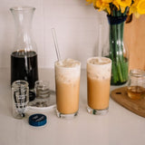 Two tall glasses of iced coffee with creamy foam, set on a countertop with a carafe, small jars, and a vase of yellow flowers in the background.