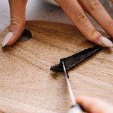 A close-up of hands slicing a dark vanilla bean on a round wooden cutting board with a small knife.