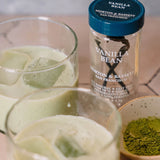 A bottle of Morton & Bassett Vanilla Bean syrup stands behind two glasses of pale green iced drink, with a small bowl of green powder in the foreground on a tabletop.