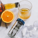 A styled flat-lay of a vanilla bean confectionery product, featuring a small glass jar of vanilla beans with a blue lid, a glass of amber liquid, a sliced orange, and sugar cubes on a white surface.