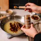 A person holds a jar of Morton & Bassett's Chinese Five Spice in one hand while sprinkling spice with a wooden spoon in another hand, with a cooking pan in the background.