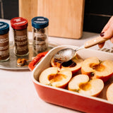 A red ceramic baking dish filled with sliced apples on a kitchen counter, with a hand using a spoon to scoop syrup. Three spice jars are visible in the background on a tray.