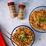 Two spice jars labeled Chinese Five Spice and Sesame Orange Ginger placed beside two bowls of stir-fried noodles with green onions, on a light gray surface with chopsticks and a red cloth.