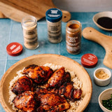 A styled food scene showing a bowl of grilled chicken and rice on a blue tiled surface, with spice jars and small bowls arranged around it. The most prominent sellable subject appears to be the spices/spice products rather than the plated meal, and the jars are clearly visible with readable labels.