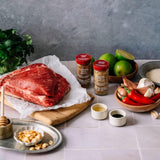 Two spice jars labeled 'Chinese Chili' and 'Garam Masala' displayed on a kitchen counter alongside raw ingredients including a cut of red beef, limes, red chilies, garlic, onions, and herbs.