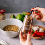 A person holds a small glass jar of Chinese Five Spice Seasoning over a kitchen counter, using a black measuring spoon to scoop the brown spice. The jar label reads 'CHINESE FIVE SPICE SEASONING' and 'BUTTER & BASSETT'.
